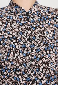 Blusa nera a fiori con colletto piccolo, piccoli fiori blu e rosa, e bottoni neri lungo la parte anteriore.