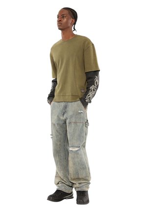 Olijfgroen shirt met korte mouwen, gedragen over een zwart shirt met lange mouwen, gecombineerd met lichtblauwe versleten wijde spijkerbroek en zwarte sneakers.