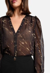 Blouse à motifs noirs et marron avec un col en V, des plis discrets et des boutons à l'avant. Associée à un pantalon noir taille haute.