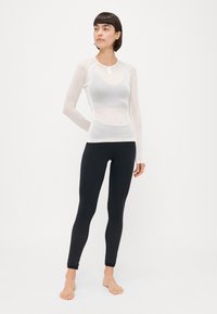 Hvid langærmet mesh træningstrøje parret med sorte tætsiddende leggings med et tekstureret mønster nær anklerne. Model står barfodet.