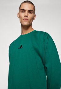 Zaļa jaciņa ar garām piedurknēm, izgatavota no kokvilnas, ar apaļu kakla izgriezumu un melnu Adidas logo uz krūtīm. Gluda tekstūra.