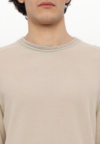 Beżowy sweter o fakturowanym splocie. Posiada wielokolorowy prążkowany kołnierz. Zbliżenie, skupiające się na dekolcie i szczegółach tkaniny.