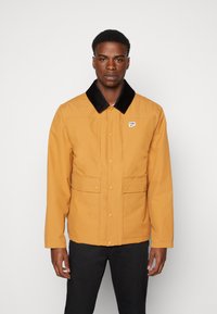 Brun zip-up jakke med en svart kordfløyelkrage, to frontlommer og en liten logo på brystet. Stoffet ser glatt og slitesterkt ut.
