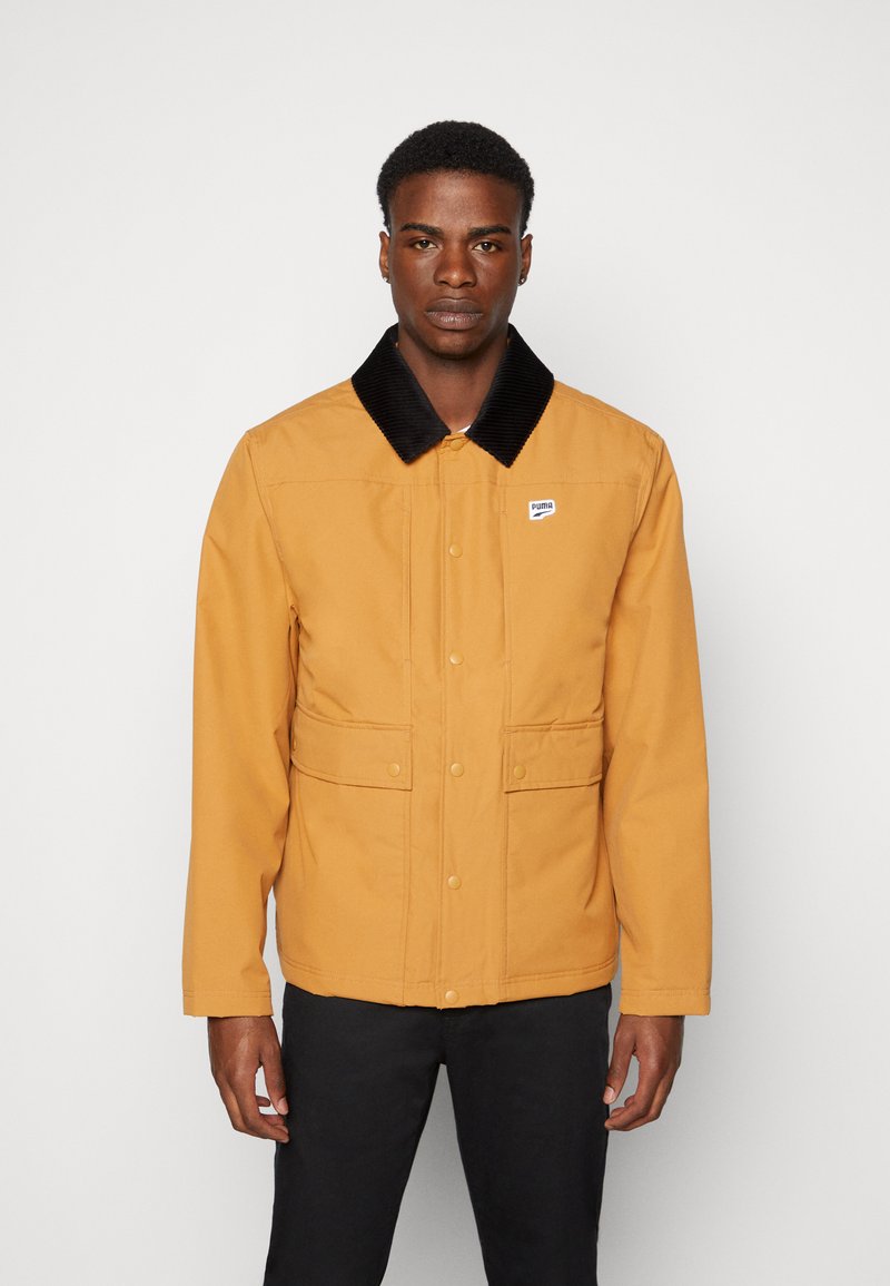 Brun zip-up jakke med en svart kordfløyelkrage, to frontlommer og en liten logo på brystet. Stoffet ser glatt og slitesterkt ut.