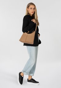 Tasche aus braunem Leder mit matter Oberfläche, ausgestattet mit doppelten Griffen und einem eleganten, kantigen Design. Das Modell trägt legere hellblaue Jeans und schwarze Sneakers.