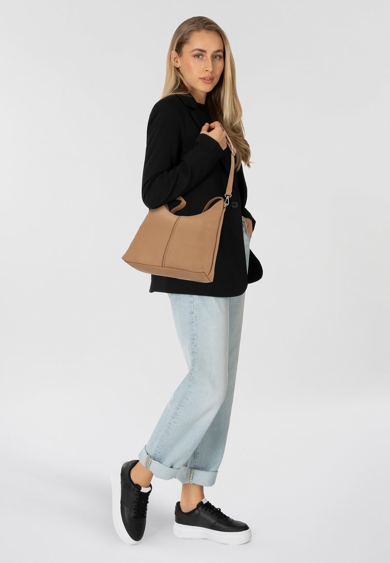 Tasche aus braunem Leder mit matter Oberfläche, ausgestattet mit doppelten Griffen und einem eleganten, kantigen Design. Das Modell trägt legere hellblaue Jeans und schwarze Sneakers.