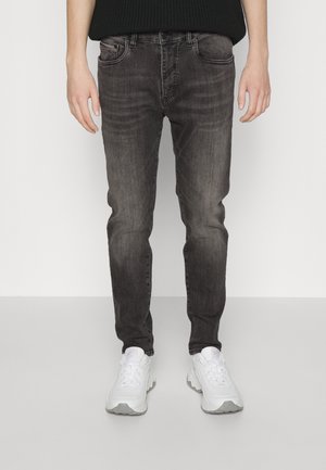 Personne portant un jean slim noir délavé et des baskets blanches épaisses, debout sur un fond clair uni.