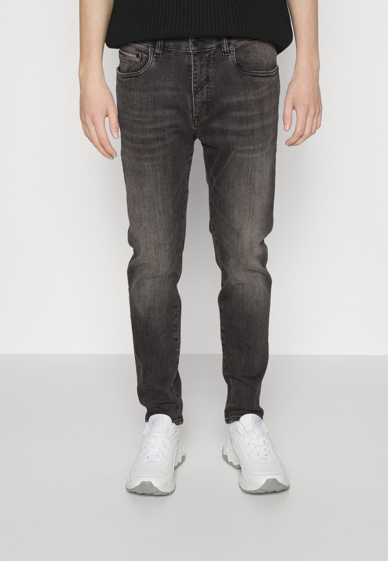 Personne portant un jean slim noir délavé et des baskets blanches épaisses, debout sur un fond clair uni.