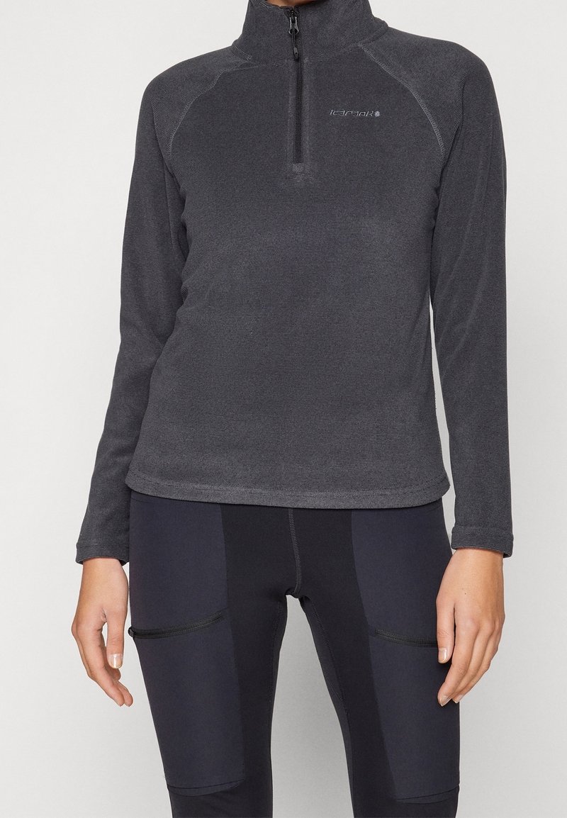 Donkergrijze fleece top met lange mouwen en rits, gemaakt van een textuuraan stof met contrasterende naden. Draagt donkere leggings met zakken met rits.