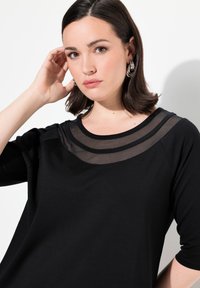 Schwarzes Oberteil mit einem transparenten Mesh-Ausschnitt, kurzen Ärmeln und einer lockeren Passform. Der Stoff hat eine glatte Textur mit subtilen Lagen-Details.