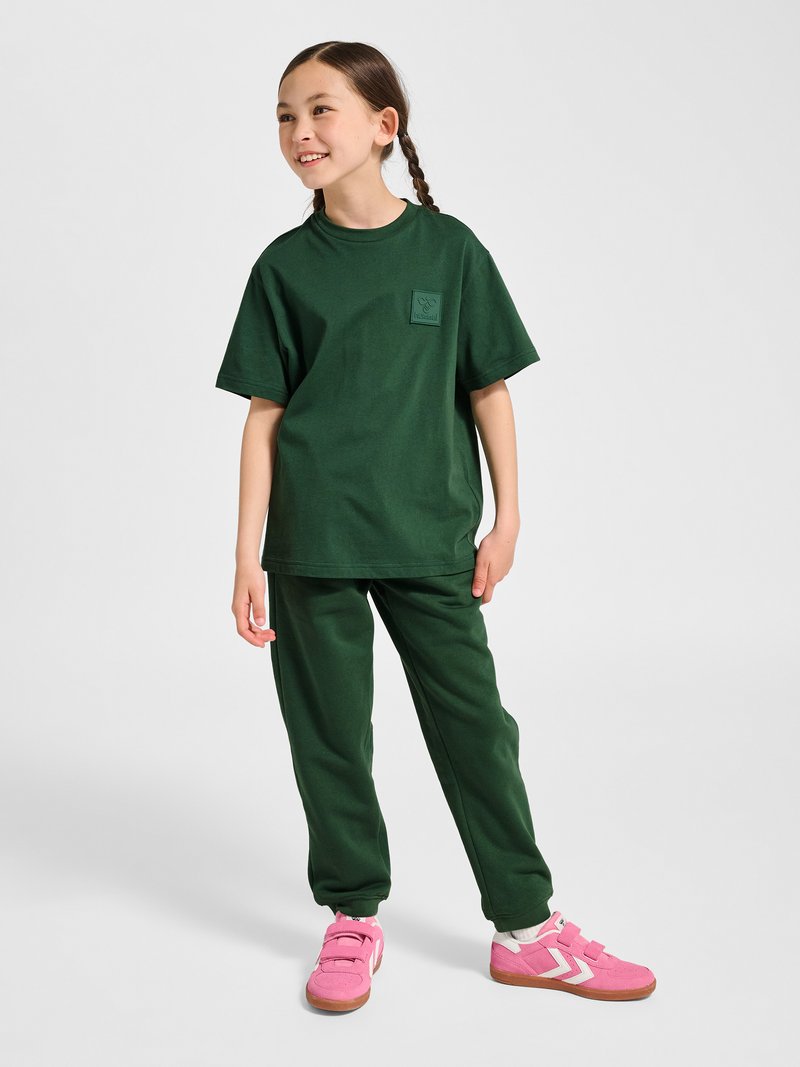 Jeune fille aux cheveux tressés portant un t-shirt ample vert foncé et un pantalon, debout et souriante, baskets roses avec des rayures blanches.
