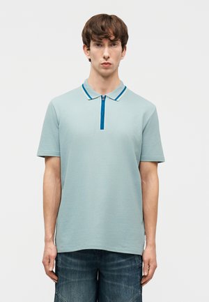 Young man standing facing forward wearing a light blue short-sleeve polo shirt with a blue zippered collar and dark blue jeans.