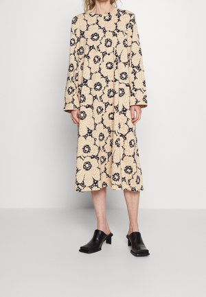 Frau trägt ein beiges Midikleid mit großen schwarz-weißen Blumenmustern und schwarzen flachen Schlupfschuhen, steht vor einem schlichten Hintergrund.