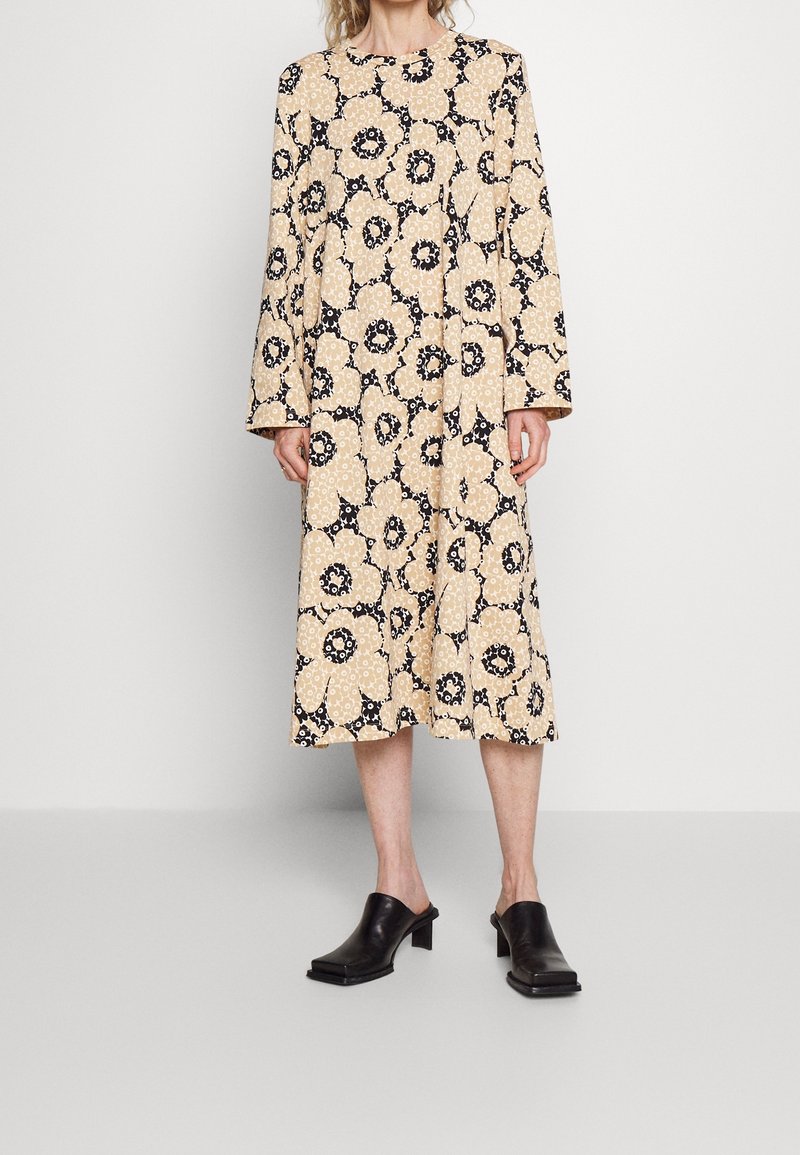 Femme portant une robe midi beige avec de grands motifs floraux noirs et blancs et des chaussures noires à talons plats, debout devant un fond uni.