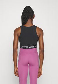 Femme aux cheveux tressés portant un haut de sport court noir et un legging rose taille haute, debout de dos sur un fond uni.