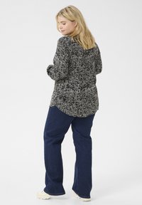 Camisa texturizada en blanco y negro con un corte holgado, con un dobladillo curvado, combinada con jeans acampanados en azul oscuro y calzado beige.