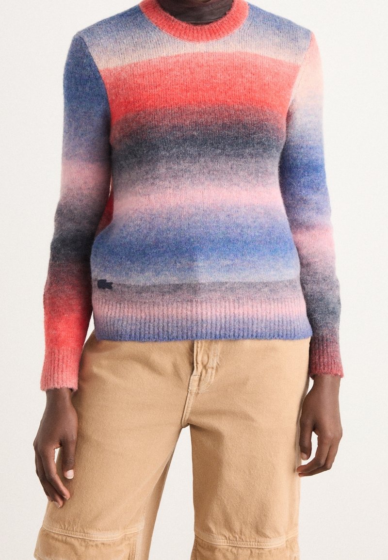 Personne portant un pull en maille à manches longues avec des rayures horizontales rouges, roses, bleues et grises, associé à un pantalon cargo beige.