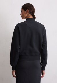 Zwarte sweatshirt met een geribde manchette en kraag; heeft een relaxed fit en een zachte textuur, weergegeven van achteren tegen een lichte muur.
