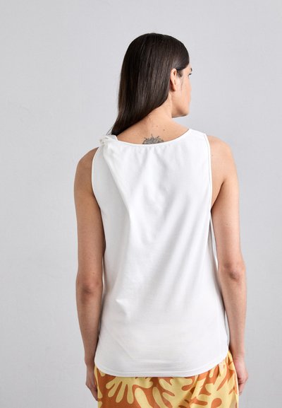 White sleeveless blouse with a relaxed fit and back tie detail, made from soft material, paired with yellow patterned skirt.