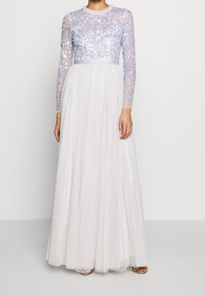 Vrouw die een lange witte jurk draagt met een doorschijnend lavendelkleurig lijfje met pailletten en lange mouwen, een vloerbedekkende tule rok en metalen open schoenen.