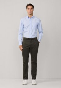 Camicia button-down a righe blu chiaro con taschino sul petto, abbinata a pantaloni verde oliva e sneakers bianche, in piedi davanti a uno sfondo neutro.