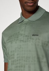 Zelena polo majica s suptilnim rešetkastim uzorkom, izrađena od meke tkanine, ima preklopljen ovratnik i crni "BOSS" logo na prsima.
