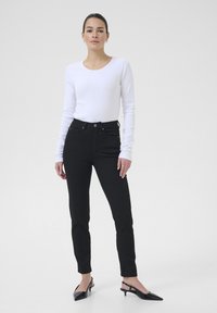 Jeans noirs taille haute à coupe slim, avec poches avant et fermeture à bouton, associés à un t-shirt à manches longues en rib de couleur blanche et des talons noirs.