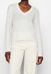 Helle beigefarbene Kabelstrickpullover mit V-Ausschnitt und langen Ärmeln, der ein strukturiertes Muster und einen gerippten Saum aufweist.