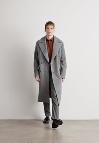 Lang grå uldfrakke med brede klapper, dobbeltbrystet front, parret med en brun polo shirt og grå bukser, båret med sorte sko.