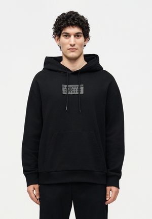 Tânăr cu păr creț poartă un hanorac negru cu buzunar frontal și un mic patch dreptunghiular cu simboluri pe piept.