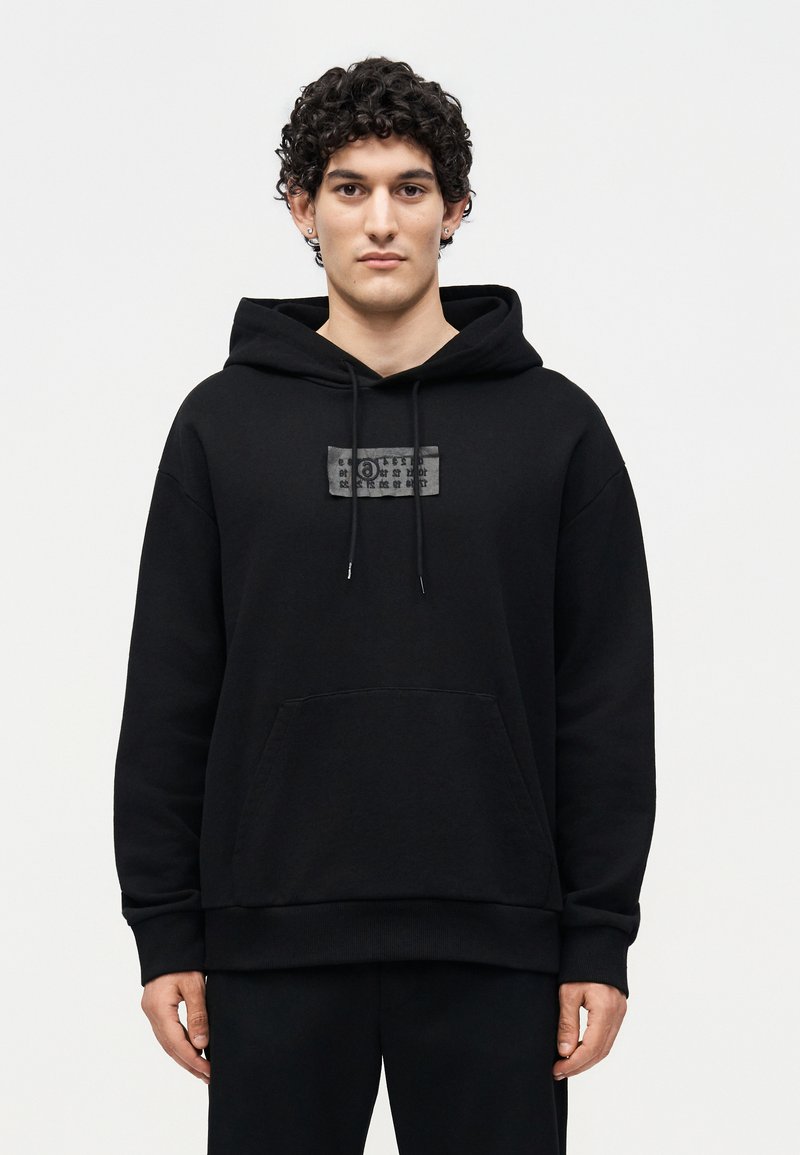 Junger Mann mit lockigem Haar trägt einen schwarzen Hoodie mit Fronttasche und einem kleinen rechteckigen Aufnäher mit Symbolen auf der Brust.