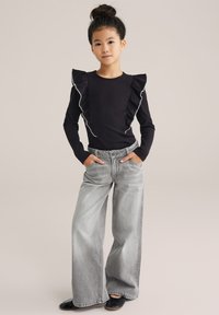 Top nero a maniche lunghe con dettagli a volant e rifinitura bianca, abbinato a jeans grigi a gamba larga. Scarpe nere completano l'outfit.