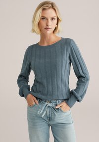 Maglione a coste azzurro chiaro con maniche lunghe, scollatura rotonda e polsini arrotolati, abbinato a jeans in denim chiaro con cintura legata in vita.