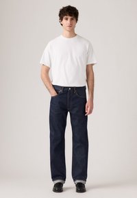 Uomo che sta in piedi contro uno sfondo neutro, indossa una t-shirt bianca a maniche corte, jeans blu scuro con risvolti e scarpe nere.