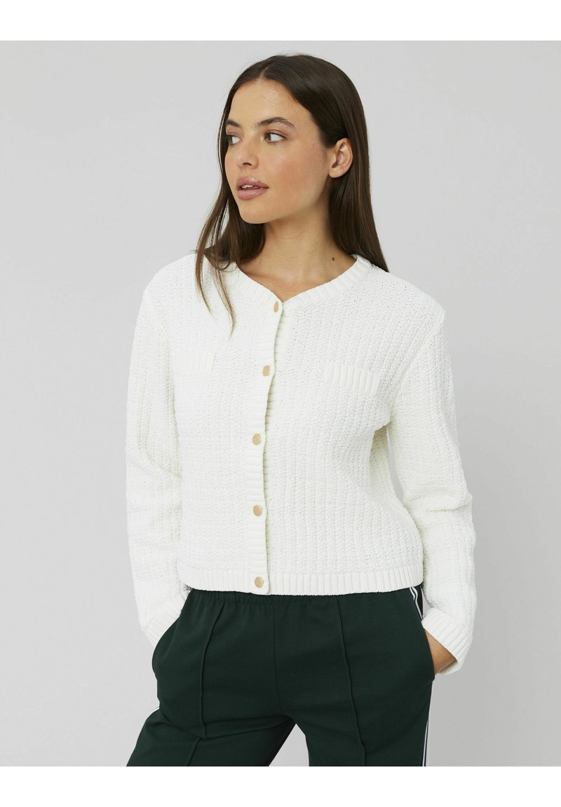 Cardigan blanc en tricot avec une texture côtelée, doté d'un col rond, de boutons en bois et de deux petites poches plaquées. Associé à un pantalon vert.