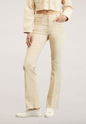 Beige high-waisted jeans met een knoopsluiting aan de voorkant, slim fit tot flared leg silhouet, met een rafelige zoom. Gecombineerd met witte sneakers.