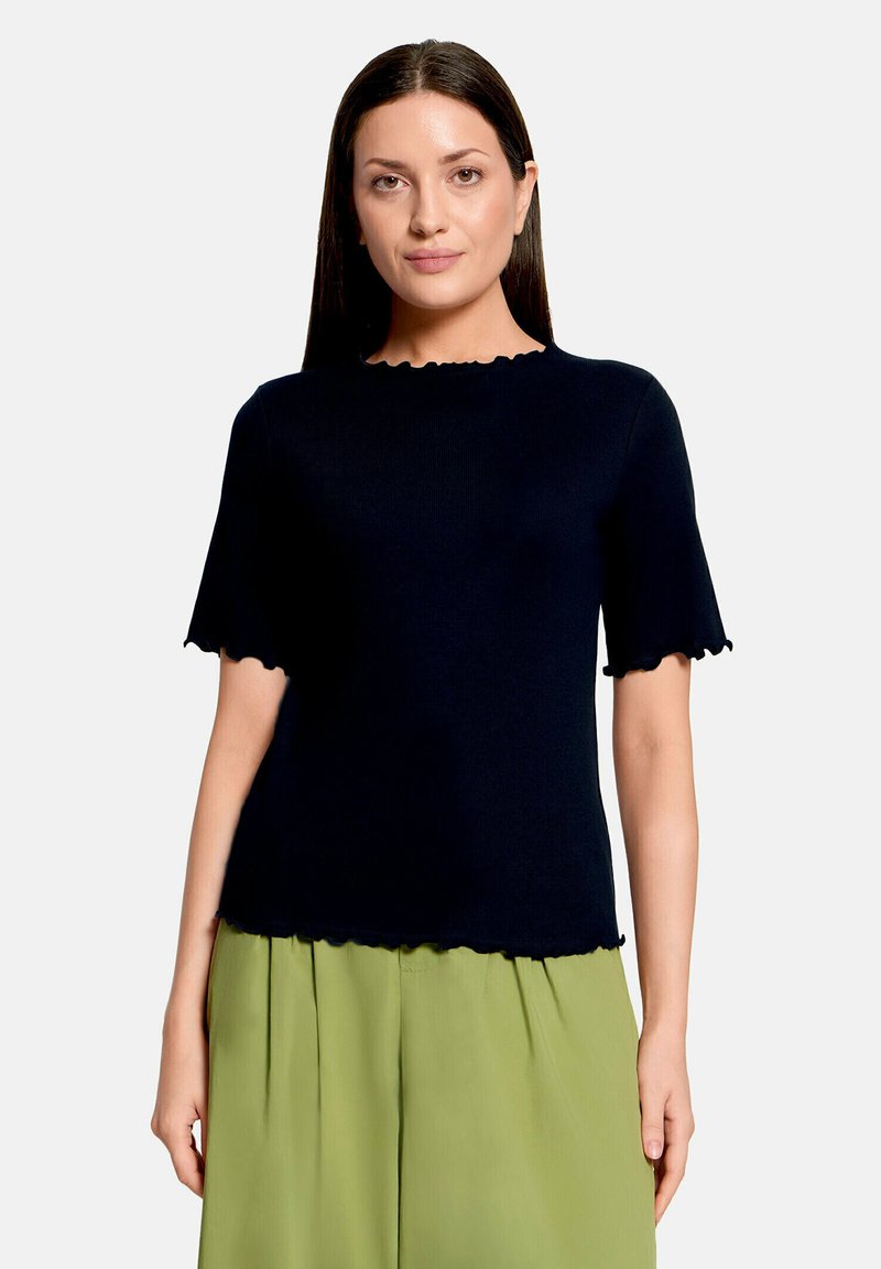 Femme aux cheveux raides et foncés, portant un haut noir à manches courtes avec des bords festonnés et un pantalon plissé vert clair, debout devant un fond blanc.