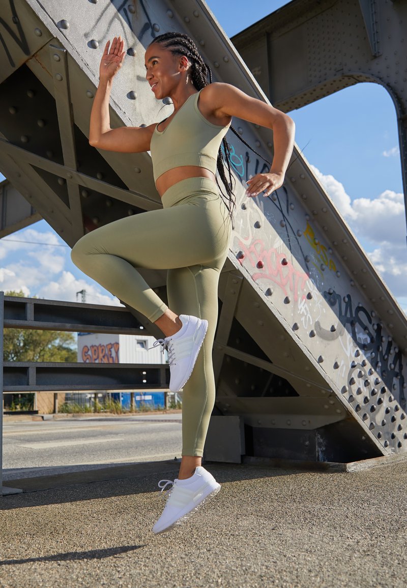 Conjunto atlético verde-azeitona com um top curto e leggings de cintura alta, combinado com sapatilhas brancas, situado perto de uma estrutura metálica.