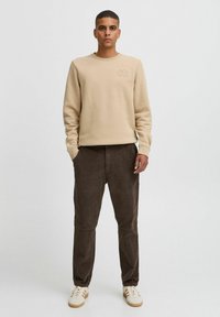 Felpa beige con logo ricamato, abbinata a pantaloni di velluto a coste marroni scuri e sneakers bianche con strisce marroni.