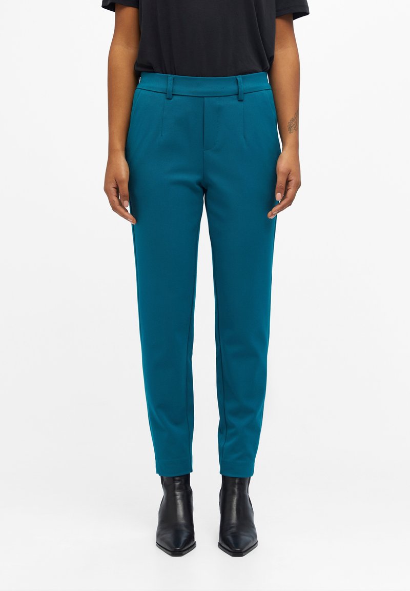Personne portant un pantalon ajusté turquoise avec passants de ceinture et des bottines noires, debout devant un fond blanc uni.