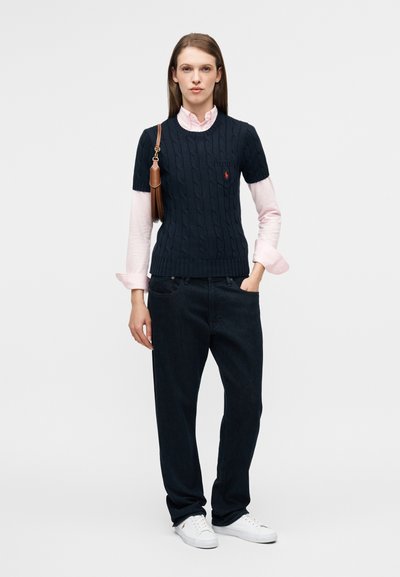 Laivastonsininen palmikkoneule, jossa on lyhyet hihat, vaaleanpunaisen pitkähihaisen paidan päällä, yhdistetty tummiin farkkuihin ja valkoisiin tennareihin. Ruskea laukku korostuksena.