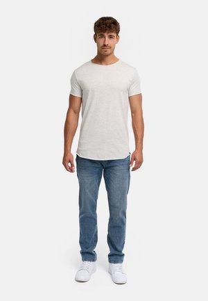 Junger Mann mit kurzen lockigen Haaren, der ein schlichtes hellgraues T-Shirt, blaue Jeans und weiße Sneakers trägt, steht vor einem weißen Hintergrund.