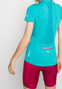 Jersey de ciclismo turquesa con mangas cortas, panel trasero de malla y bolsillo trasero. Combinado con pantalones cortos acolchados color burdeos, de textura suave.