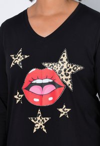 Musta V-kauluspaita, jossa on punainen huuligrafiikka, vaaleanpunaisia yksityiskohtia ja kolme beigeä leopardikuvioista tähteä, joissa on strassiyksityiskohtia.