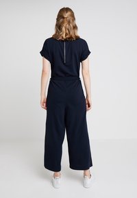 Žena s vlasy po ramena staženými vzadu, oblečená v námořnické modré kombinéze s ohrnutými rukávy a bílými teniskami, stojící před jednobarevným pozadím.