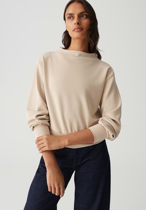 Sweatshirt beige à manches longues avec un col montant, des poignets côtelés et un ourlet élastique. Associé à un jean foncé ; texture de tissu lisse.