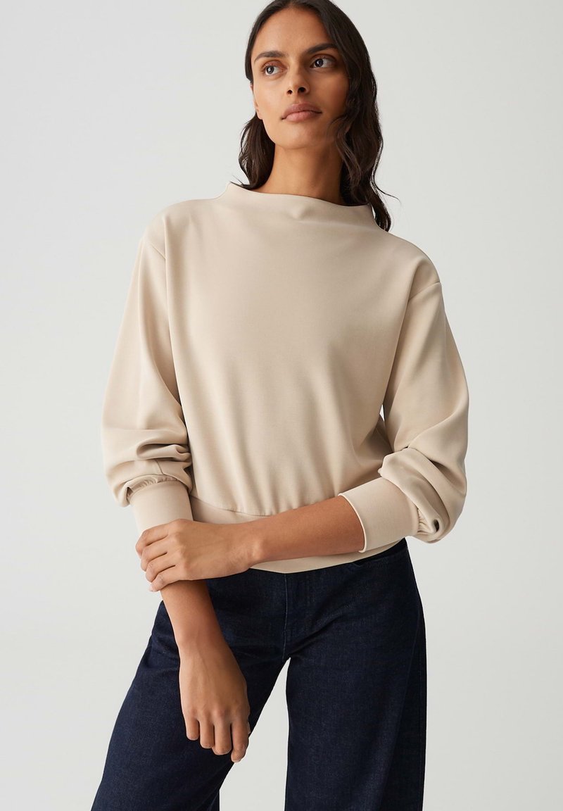 Felpa beige a maniche lunghe con colletto alto, polsini a coste e orlo elastico. Abbinata a jeans scuri; texture del tessuto liscia.