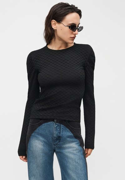 Γυναίκα που φοράει μαύρο υφασμάτινο τοπ με μακριά μανίκια και τζιν δύο αποχρώσεων μπλε και μαύρου, με μικρά μαύρα γυαλιά ηλίου, πόζαρε μπροστά σε λευκό φόντο.