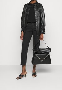 Černá kožená taška s přehnutým zavíráním a řetízkovým uchem. Modelka má na sobě černou koženou košili, černý top a zkrácené černé džíny, s otevřenými lodičkami.