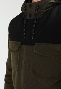 Kurtka oliwkowa z czarną górną częścią, z kapturem, dwoma kieszeniami na piersi i regulowanymi sznurkami. Teksturowane wykończenie tkaniny.